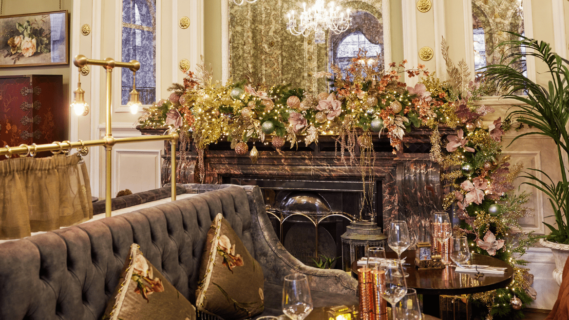 Elegant lounge seating area with a marble fireplace decorated with festive greenery, ornaments, and warm fairy lights, surrounded by plush sofas and set tables with glassware.