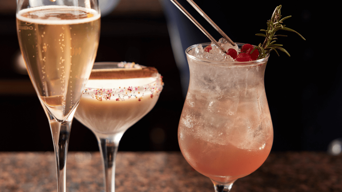 Assorted cocktails on a bar counter, including a champagne flute, a creamy cocktail with sprinkles, and a pink iced drink garnished with berries and rosemary.
