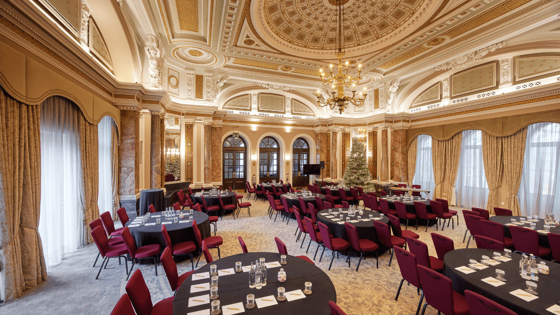 Grand ballroom with high ornate ceilings, chandeliers, and round tables set with black linens and red chairs, arranged for a formal event or conference.