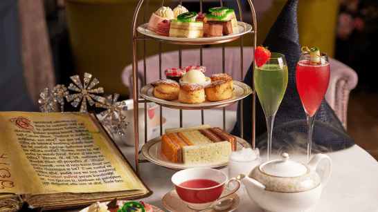 Wicked three-tier afternoon tea display featuring cakes, scones, and finger sandwiches, served alongside a teapot, teacup, and two brightly colored cocktails. A decorative spellbook and themed ornaments create a whimsical, magical atmosphere.