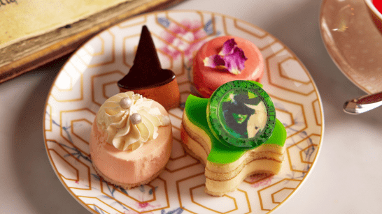 Close-up of a patterned dessert plate featuring a pink mousse topped with piped cream, a green star-shaped layered cake with a printed chocolate disc, a pink glazed pastry topped with a flower petal, and a small chocolate cone-shaped dessert.