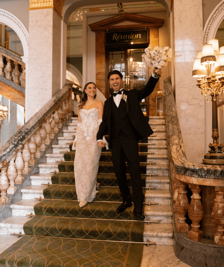 A couple in formal wedding attire walking down the grand marble staircase inside the Clermont Victoria, holding hands, with one person holding a bouquet, ornate balustrades, chandeliers, and the entrance to “Réunion” visible in the background.