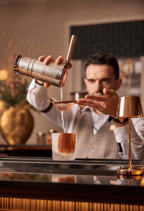 Bartender pouring an amber drink from a shaker into a glass using a strainer, with a gold lamp and floral vase in the background