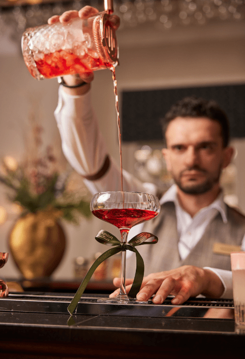 Bartender pouring a red cocktail into a coupe glass with a green ribbon, with a gold vase and flowers in the background