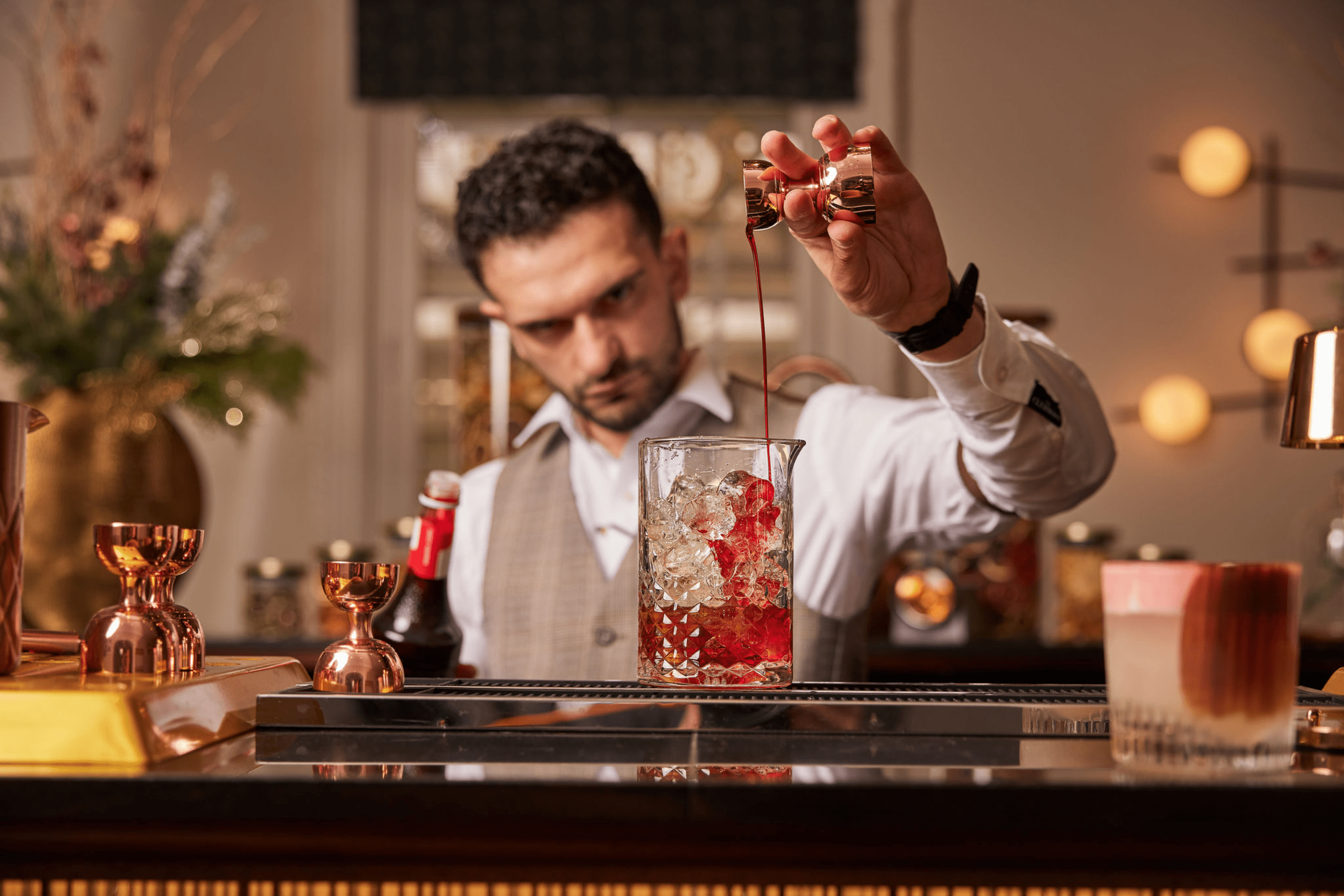 Bartender pouring red liquid into a cocktail glass with ice, surrounded by bar tools and decorations