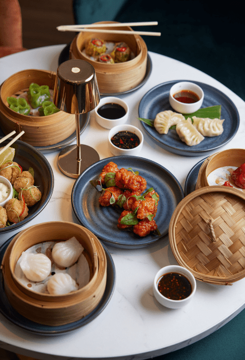 Assorted dim sum dishes on a round white table with bamboo steamers, blue plates, dipping sauces, and a small lamp in the center