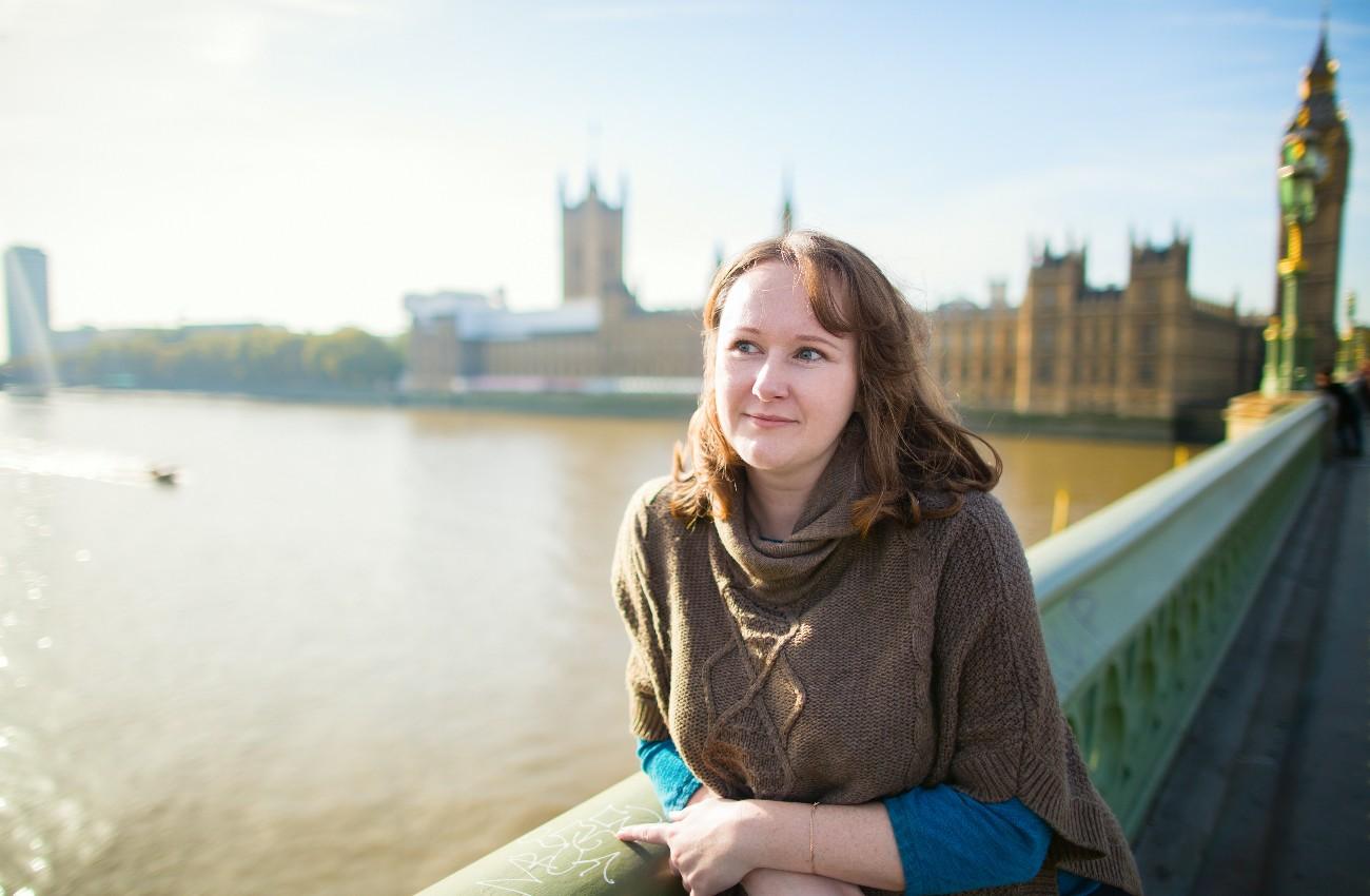 Woman standing on Westminster Bridge beside the River Thames, with the Houses of Parliament and Big Ben softly blurred in the background on a bright day.
