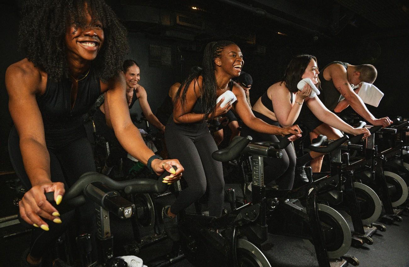 Group of people taking part in an energetic indoor spin class, riding stationary bikes and smiling during the workout.