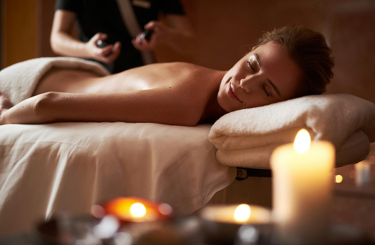 Woman relaxing face down on a massage table in a warmly lit spa, with folded towels and glowing candles in the foreground.