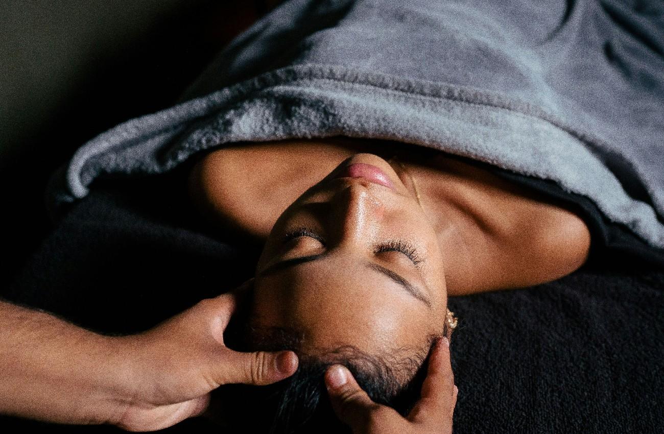 Woman with her eyes closed receiving a scalp massage, covered with a grey towel.