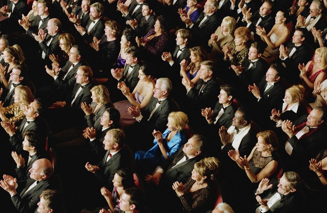 Theatre audience in formal evening wear applauding from rows of seats.