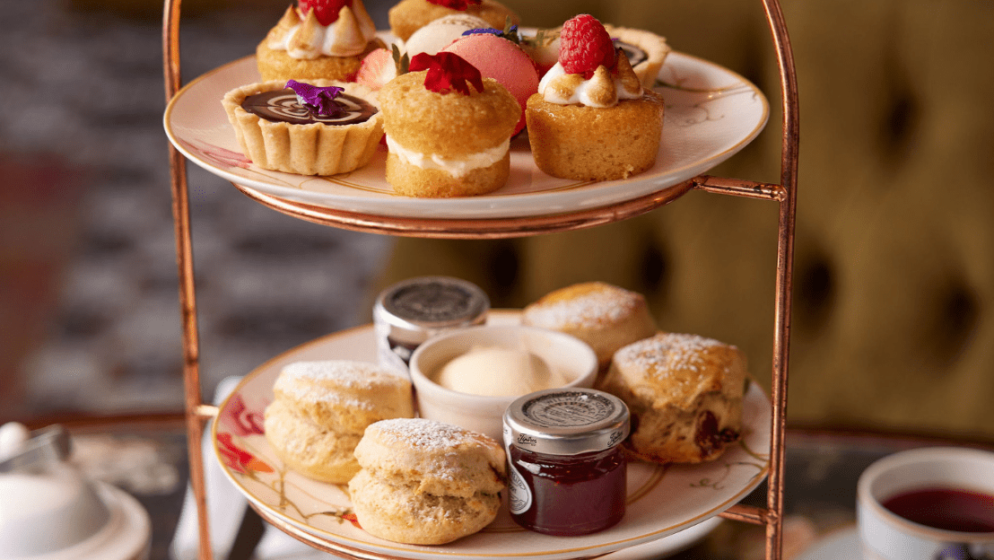 Two-tiered stand with assorted pastries, scones, and a cup of tea in the background