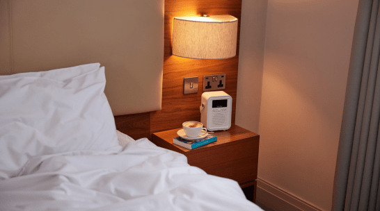 Bedside table with a lamp, alarm clock, books, and a cup beside a neatly made bed